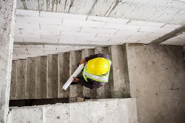 Trabajador joven ingeniero profesional en casco protector y papel de planos en mano en el sitio de construcción de edificios de viviendas