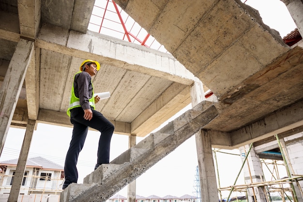 Trabajador joven ingeniero profesional en casco protector y papel de planos en mano en el sitio de construcción de edificios de viviendas