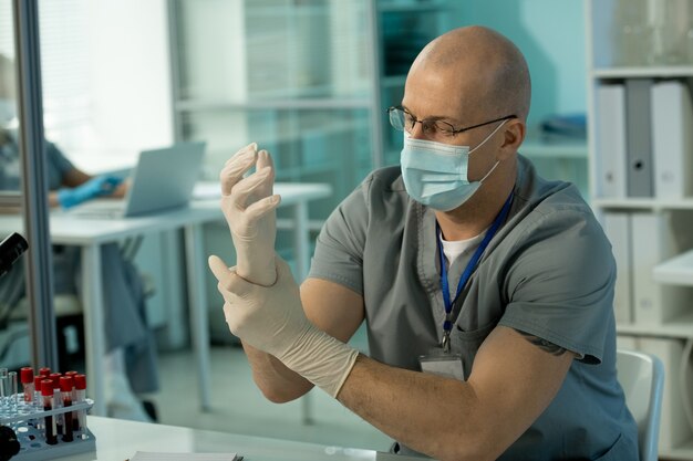 Trabajador de laboratorio calvo en máscara sentado a la mesa y ponerse guantes mientras se prepara para la investigación de análisis de sangre