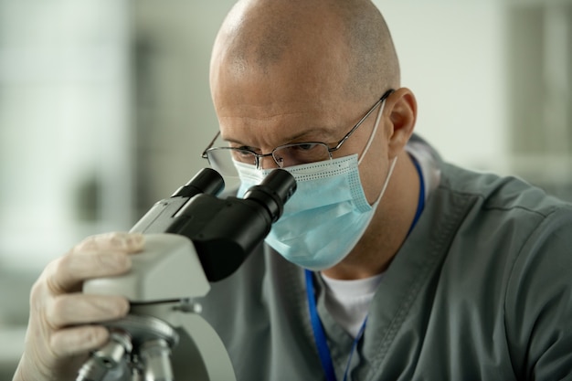 Trabajador de laboratorio concentrado en gafas y máscara con microscopio mientras analiza la muestra en el laboratorio