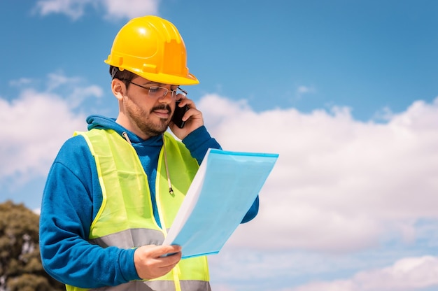 Trabajador latino con casco amarillo, gafas y chaleco verde, hablando por teléfono y sosteniendo una carpeta en la mano