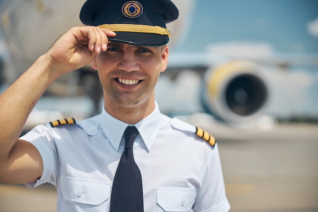 Trabajador de línea aérea joven alegre en gorra piloto de pie al aire libre en el aeropuerto