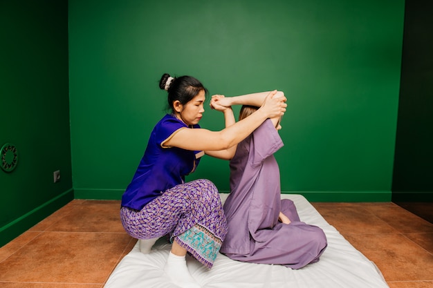 Trabajador masajista tailandés en ropas étnicas asiáticas hace procedimientos de spa tradicionales a la bella dama blanca en pijama morado en la sala de yoga verde