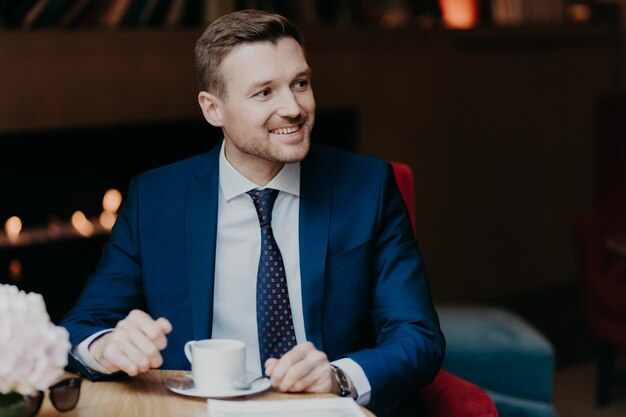 El trabajador masculino alegre en ropa formal mira felizmente a un lado, bebe café aromático caliente, feliz de notar a alguien a un lado