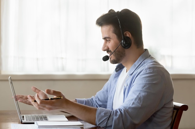 Trabajador masculino con auriculares consulta a los clientes en línea en la computadora portátil