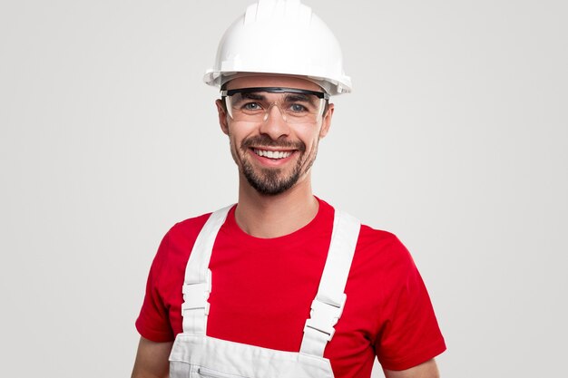 Trabajador masculino barbudo amistoso positivo en casco y gafas generales y protectores sonriendo y mirando a la cámara contra el fondo blanco