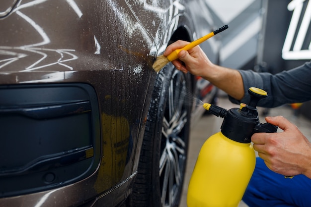 El trabajador masculino limpia la superficie del automóvil con spray y cepillo, preparación antes de aplicar la película protectora, detallando. Instalación de revestimiento que protege la pintura de automóvil de arañazos.