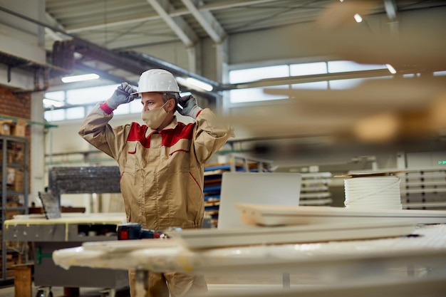 Trabajador masculino con mascarilla usando casco de trabajo mientras trabaja en el taller de carpintería