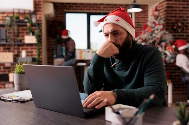 Trabajador masculino que usa una computadora portátil en la oficina comercial para trabajar en un proyecto de inicio durante la temporada de invierno. Hombre con gorro de Papá Noel trabajando en un informe en el lugar de trabajo con árbol de Navidad y adornos navideños.