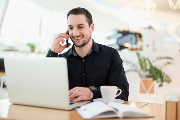 Trabajador masculino remoto exitoso. Hombre joven autónomo feliz con una llamada telefónica. Cafetería en el fondo. Programador autónomo masculino que trabaja en una cafetería. Él está feliz de hablar con el cliente.