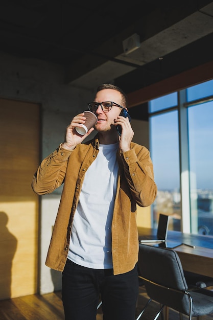 Trabajador masculino sonriente con ropa informal mirando hacia otro lado mientras habla por teléfono móvil y bebe café durante el descanso en el espacio de trabajo creativo moderno