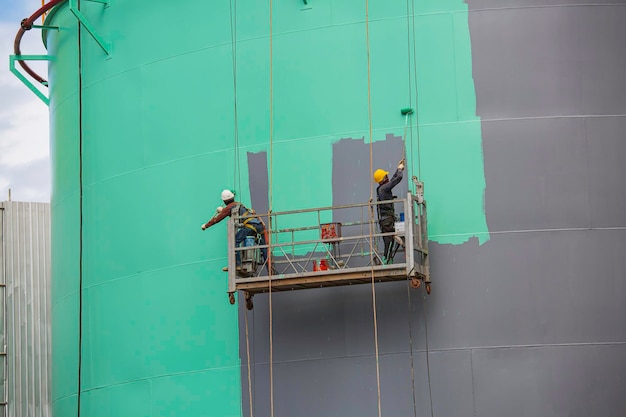 El trabajador masculino toma el teleférico sosteniendo un pincel de pintura industrial utilizado para la superficie de la superficie del aceite del tanque de la placa de la carcasa de color verde