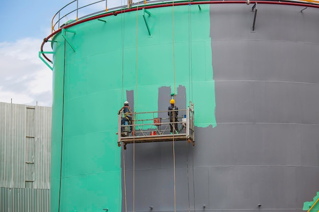 El trabajador masculino toma el teleférico sosteniendo un pincel de pintura industrial utilizado para la superficie de la superficie del aceite del tanque de la placa de la carcasa de color verde