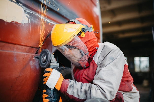 trabajador masculino en un traje con un molinillo de ángulo y un casco haciendo reparación de carrocería