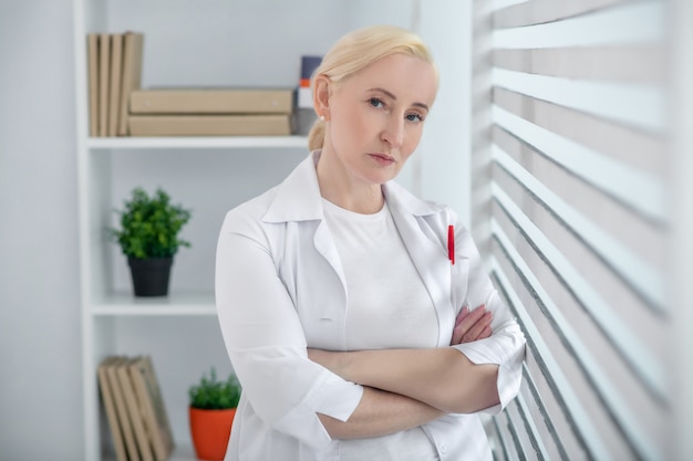 Trabajador médico Grave mujer rubia en bata blanca médica brazos cruzados de pie cerca de una ventana con persianas.