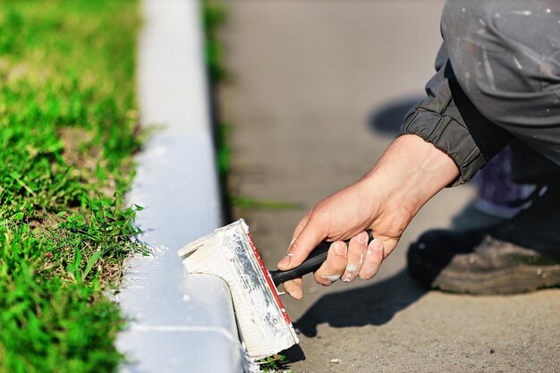 Un trabajador con un mono pinta un borde con pintura blanca en un día de verano Paisajismo de servicios urbanos