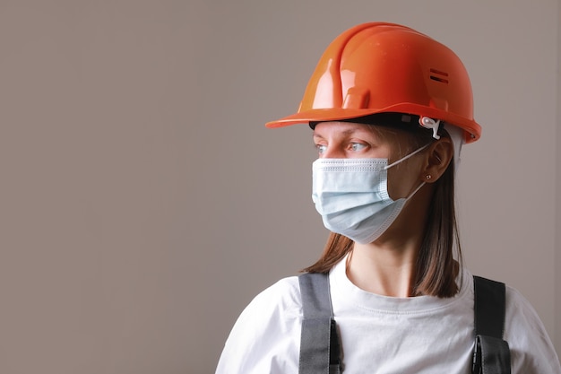 Foto trabajador de mujer bonita en máscara médica y casco de construcción naranja sobre un fondo gris.