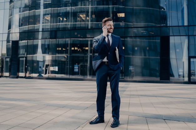Trabajador de oficina barbudo joven hermoso en traje formal azul oscuro habla por teléfono celular mientras está parado afuera al lado del centro de negocios esperando que los socios se reúnan. Concepto de gente de negocios