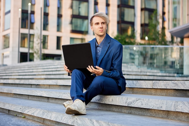 Un trabajador de oficina con una computadora portátil de rodillas se sienta en el paso frente al edificio del centro de negocios