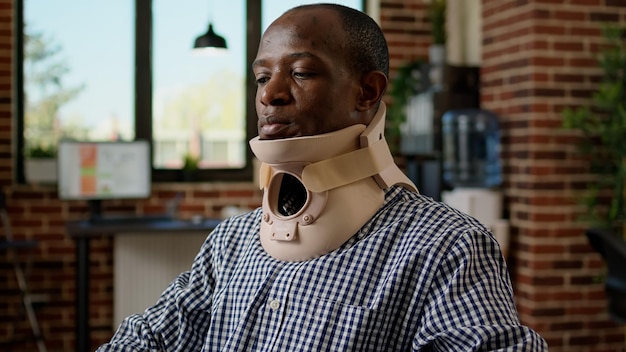 Trabajador de oficina con dolor trabajando con espuma de cuello médico en el escritorio, sufriendo un doloroso accidente por lesiones. Usar collar cervical para recuperarse del dolor de vértebras de la columna, sentirse cansado y enfermo.