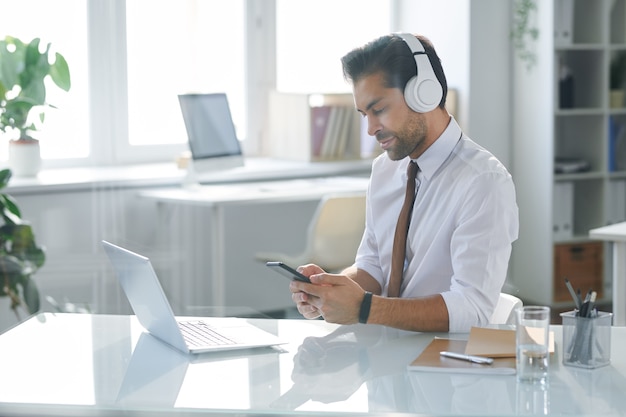 Trabajador de oficina elegante en auriculares sentado junto al escritorio mientras busca banda sonora en el teléfono inteligente