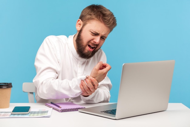 Trabajador de oficina de hombre barbudo en camisa blanca haciendo muecas sintiendo hormigueo entumecimiento y dolor en la muñeca trabajando en la computadora portátil que sufre el síndrome del túnel carpiano Disparo de estudio interior aislado sobre fondo azul