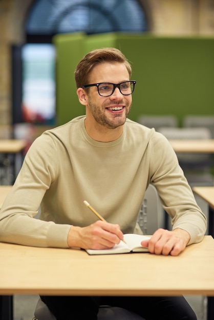 Trabajador de oficina masculino alegre sentado en el escritorio en el espacio de coworking haciendo algunas notas y sonriendo