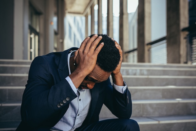 Trabajador de oficina masculino cansado después del trabajo, afroamericano tiene dolor de cabeza severo, sentado en las escaleras cerca de la oficina