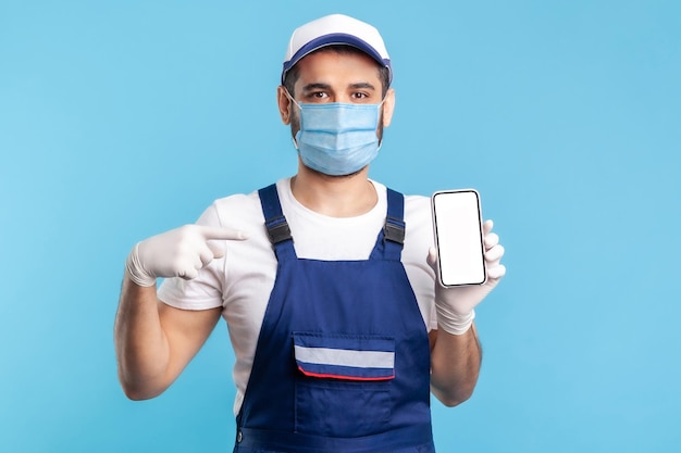 Trabajador en overoles, máscara y guantes apuntando al teléfono móvil con una pantalla en blanco simulada, área de publicidad para la aplicación de pedidos de entrega en línea, servicios de mantenimiento de reparación de casas. tiro interior aislado en azul