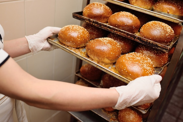 Trabajador de panadería mujer sosteniendo una bandeja de bollos redondos frescos para hamburguesas con primer plano de semillas de sésamo