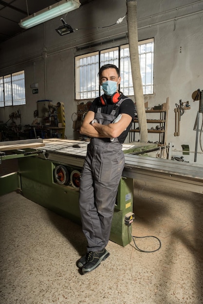 Trabajador de pie con los brazos cruzados vistiendo mascarilla y equipo de protección mirando a la cámara sonriendo en una fábrica. Tiro largo, cuerpo completo.