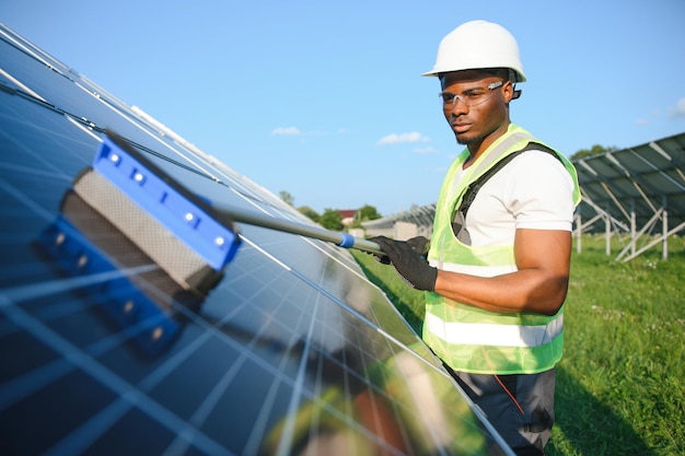 Trabajador de una planta de energía alternativa en uniforme limpiando paneles solares con una escoba Africano-Americano guapo cuidando del equipo