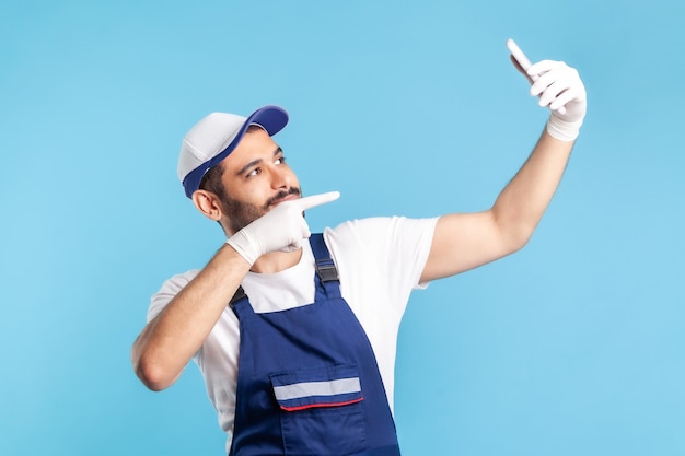 Trabajador positivo en overoles y guantes señalando con el dedo la videollamada de teléfono móvil, comunicándose en línea con el cliente, aceptando pedidos como servicios de entrega, reparación y mantenimiento. tiro del estudio aislado