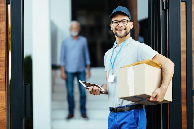 Trabajador postal feliz con tableta digital llevando paquetes y mirando la cámara mientras realiza la entrega a domicilio