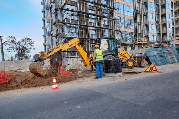 Foto trabajador profesional con ropa protectora y casco naranja está de pie cerca de una excavadora contra un moderno edificio de gran altura en construcción