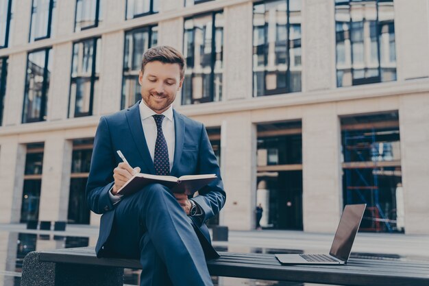 Trabajador profesional sonriente de la empresa en traje formal escribiendo ideas con bolígrafo en el cuaderno mientras está sentado en un banco con las piernas cruzadas, computadora portátil abierta junto a él, edificios de oficinas en el fondo borroso