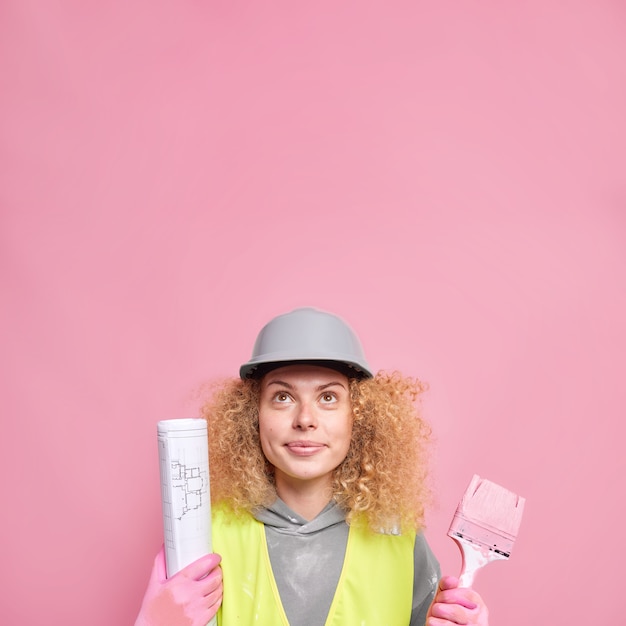 El trabajador de reparaciones del hogar tiene el cabello rizado enfocado arriba sostiene un pincel de pintura y un plano de papel ocupado con la decoración de la habitación del apartamento usa poses uniformes de casco