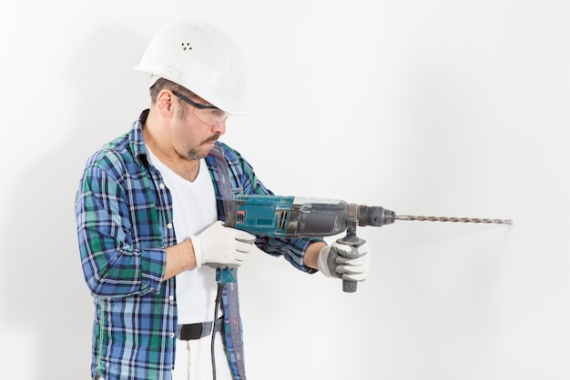 Trabajador reparador, con casco protector y gafas, perfora una pared blanca con un perforador