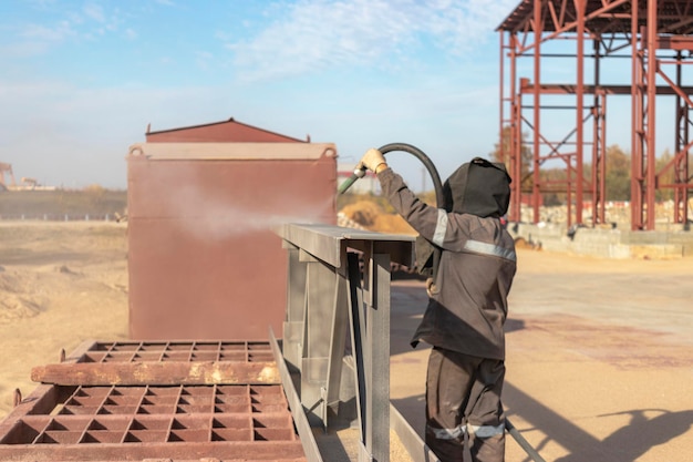 Un trabajador con ropa especial está limpiando con chorro de arena una estructura de construcción de metal en un sitio industrial Procesamiento industrial de metales