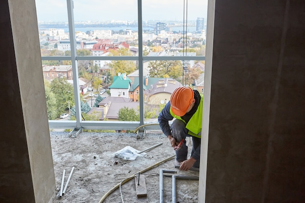 Trabajador en ropa protectora y casco de seguridad instalando tubos de plástico con herramientas modernas en el piso del edificio en construcción