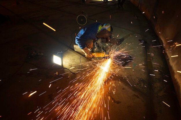 Trabajador con rueda eléctrica rectificado por chispa en la placa inferior de la pieza de acero al carbono de metal del soldador dentro del espacio confinado del tanque