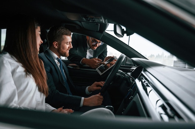 El trabajador de la sala de exposición de automóviles está consultando al hombre y la mujer que están sentados dentro del automóvil