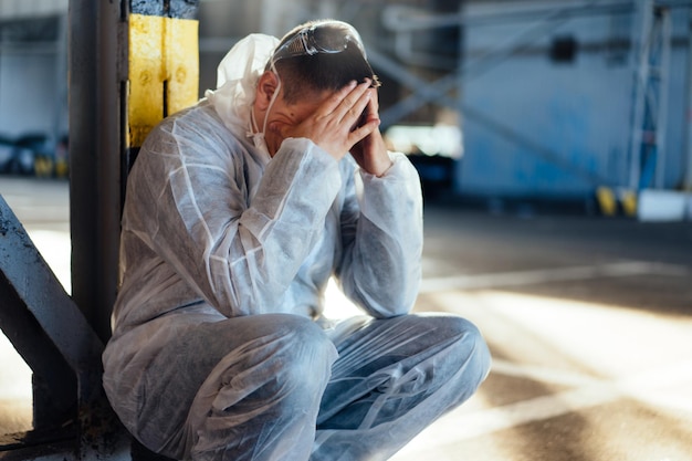 Trabajador de la salud desesperado con overoles protectores covid blancos. Cansado El médico caucásico masculino agarra su cabeza encogido en depresión durante la pandemia de coronavirus Paramédico con exceso de trabajo llanto cubierto de cara con las manos