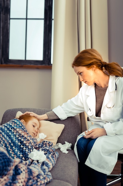 Trabajador de la salud. Doctora profesional sentada cerca de su paciente mientras toca su frente