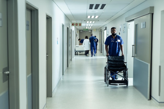 Trabajador sanitario birracial empujando una silla de ruedas vacía en un concurrido pasillo del hospital