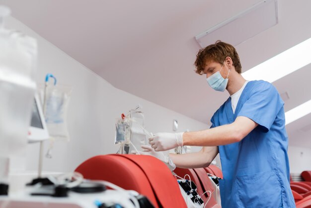 trabajador sanitario pelirrojo con máscara médica y guantes de látex montando un conjunto de transfusiones cerca de automatizado