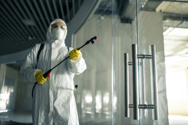 Un trabajador sanitario rocía un centro de negocios vacío con líquido antiséptico para prevenir la propagación del covid-19. Un hombre vestido con traje de desinfección limpiando el centro comercial. Nadie, salud, concepto aislado.