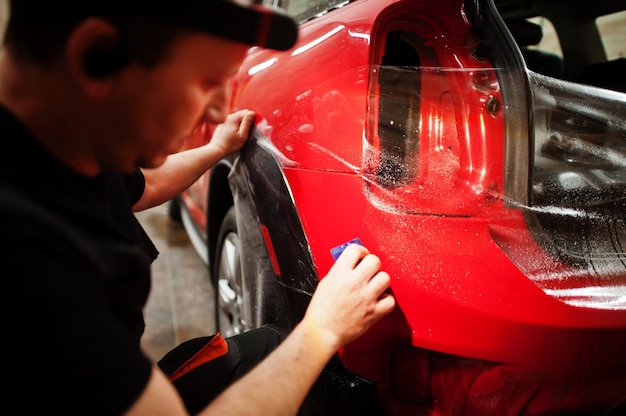 El trabajador del servicio de automóviles puso una película anti-grava en la carrocería de un automóvil rojo en el taller de detalle del vehículo