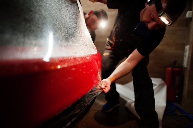 Foto el trabajador del servicio de automóviles puso una película anti-grava en la carrocería de un automóvil rojo en el taller de detalle del vehículo