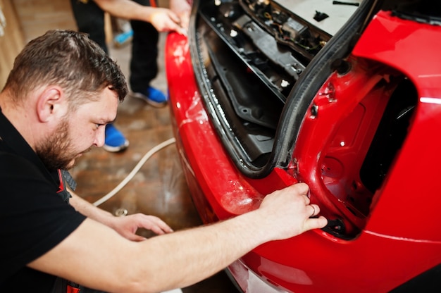 Foto el trabajador del servicio de automóviles puso una película anti-grava en la carrocería de un automóvil rojo en el taller de detalle del vehículo